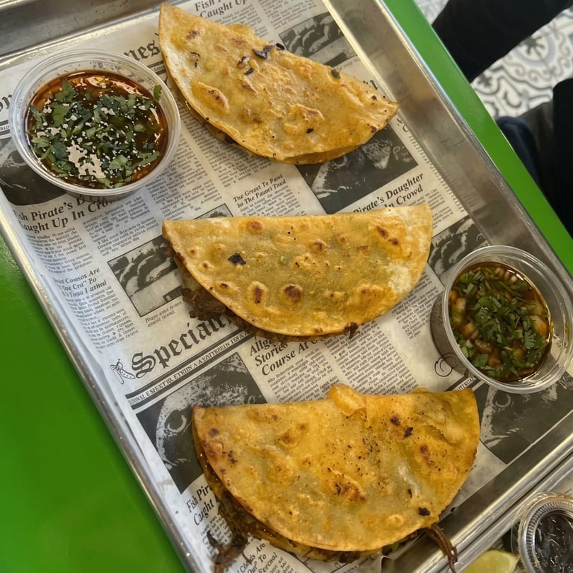 Three golden-brown quesadillas with two small cups of dipping sauce on a metal tray lined with newspaper.