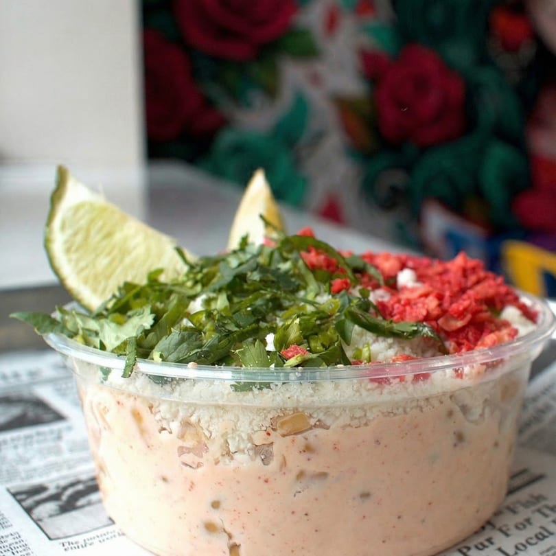 Elote in a clear cup, topped with cilantro, red spice, cheese, and a lime wedge.