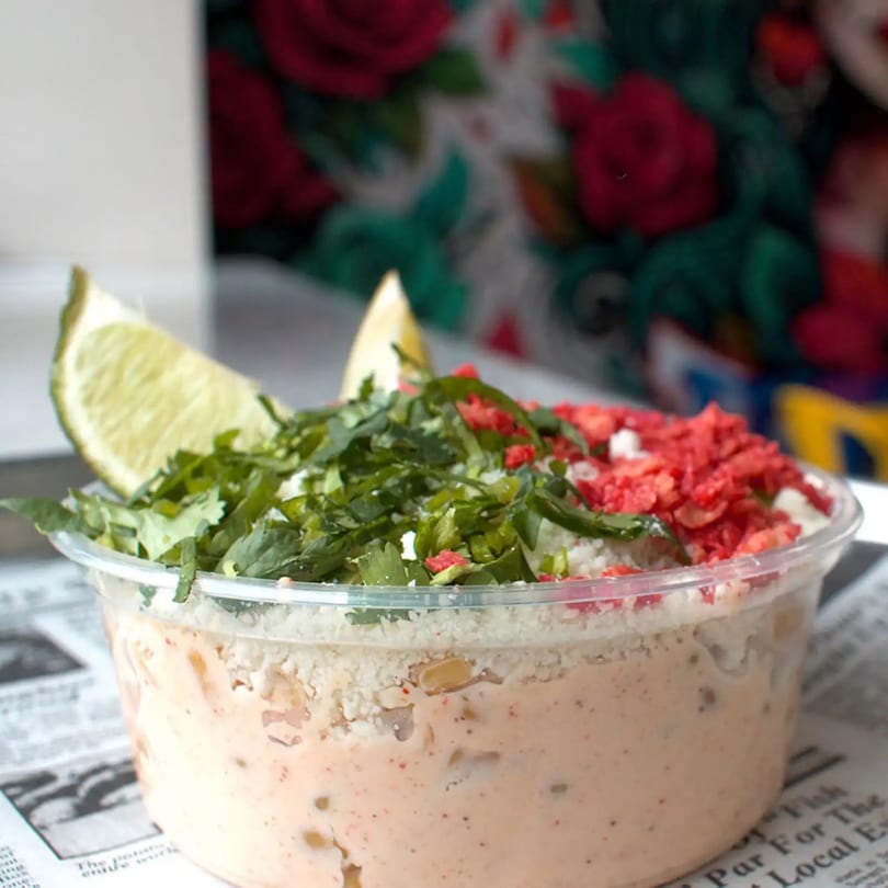 Mexican street corn in a clear cup, topped with cilantro, red chili flakes, and a lime wedge.