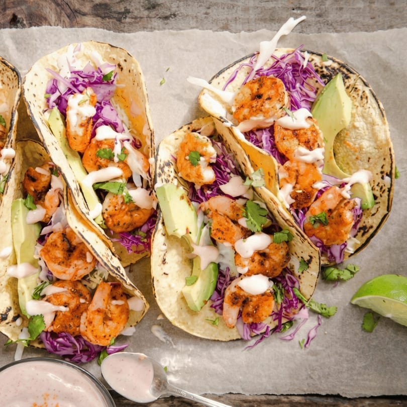 Three shrimp tacos with red cabbage, avocado, and white sauce on parchment paper.