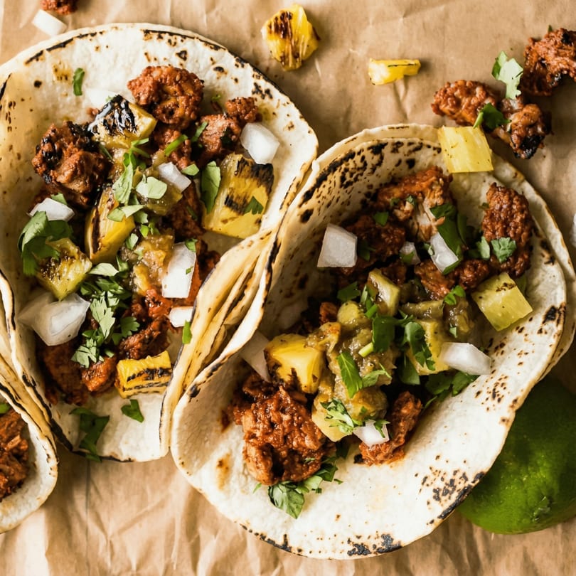 Three al pastor tacos with grilled pineapple, onions, and cilantro on a brown paper background.